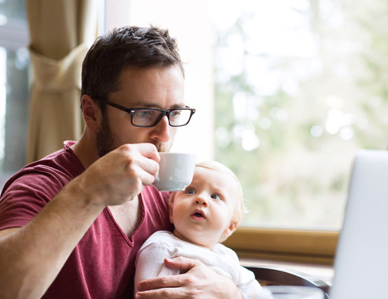 Hombre sentado y sujetando a una bebé con un brazo mientras toma café y trabaja