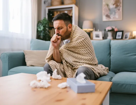 Hombre enfermo, abrigado con una manta y tosiendo en el salón de su casa